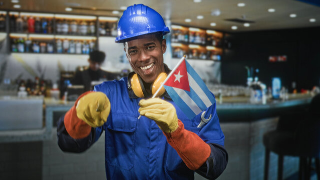 Man pointing finger and holding cuban flag, wearing blue hardhat, yellow gloves and ear protectors inside building bar; pride solidarity.