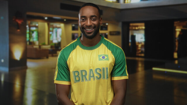 Man smiling, standing in a hotel building lobby wearing a yellow and green brazil jersey with flag patch, facing camera; pride.