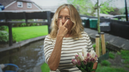 Fototapeta premium Senior blonde woman holding a bouquet of pink tulips points index finger toward camera in street by canal and house wearing a striped shirt and smiling; amused curiosity.