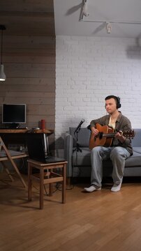 Vertical video man wearing headphones and casual clothing sitting on a couch, playing an acoustic guitar and singing into a microphone, recording music with a laptop in a home studio