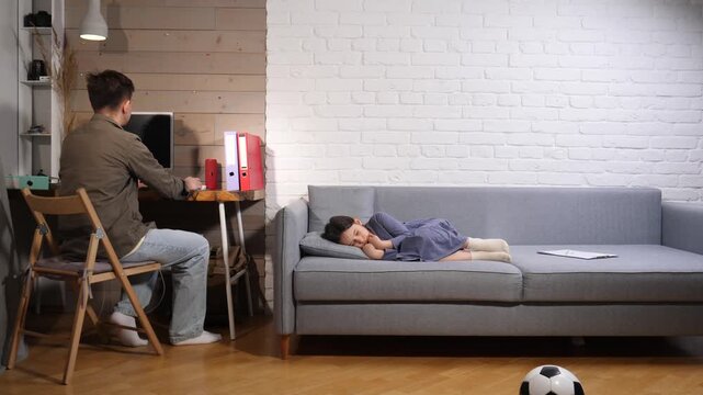 Father focused on laptop at desk in a busy home office while his young daughter naps on the sofa nearby, capturing the reality of remote work and parenting balance