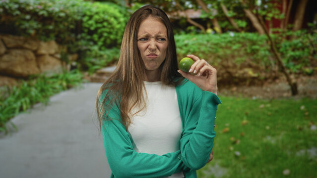 Woman in green cardigan holding a lime up to her nose on a forest path with crossed arms and a disgusted expression; disgust aversion distaste.