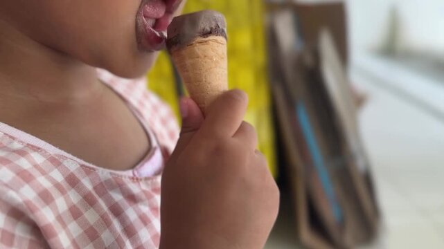 Close up of a cute little girl in a checkered dress enjoying a chocolate ice cream cone with a messy face.