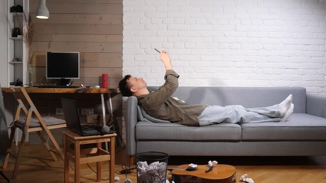 Man on a couch with an acoustic guitar, pen and notebook, surrounded by crumpled papers, deep in thought during a songwriting block in a home studio setting