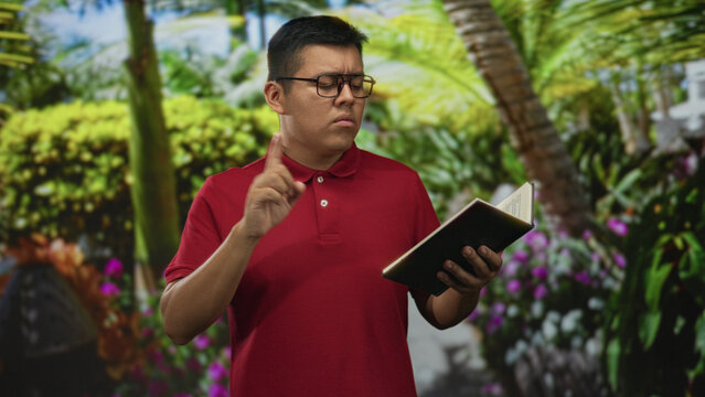 Young hispanic man wearing glasses points index finger while holding an open book in a green park with palm trees and shrubs; quiet concentration.