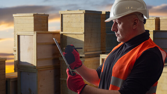 Warehouse logistics, Industrial inspection. Worker in hard hat and safety vest checks inventory on clipboard among stacked wooden crates at storage site.