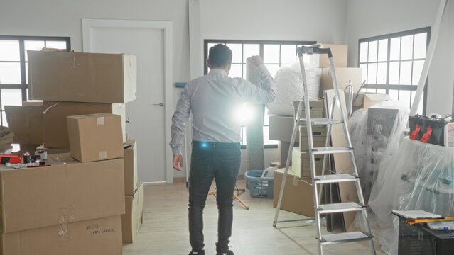 Man surrounded by stacked cardboard moving boxes, a metal ladder and covered furniture raises arms while facing a closed door inside a building entryway; fresh start relief.