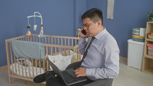 Man holds smartphone to ear and types on laptop beside a cradle in a baby room building; work life stress.