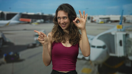 Obraz premium Woman making ok sign with hand in front of airplane at busy airport terminal during daytime; confidence.