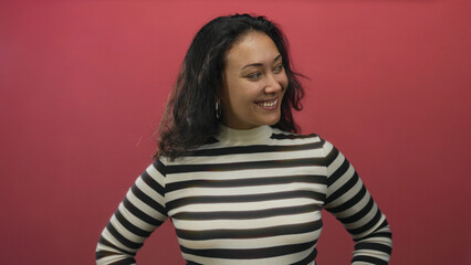 Fototapeta premium Young hispanic woman smiling with hands on hips in red studio wearing black and white striped turtleneck and hoop earrings; confidence.