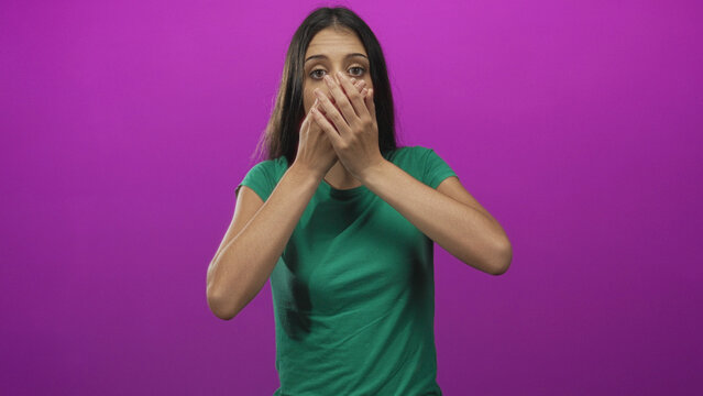 Young woman in green shirt with hands covering mouth, wide eyes and visible gasp in purple studio; surprise concern.