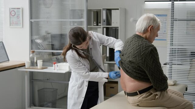 Young adult female medical professional placing stethoscope on back of elderly man while listening to lung sounds during physical exam in medical office