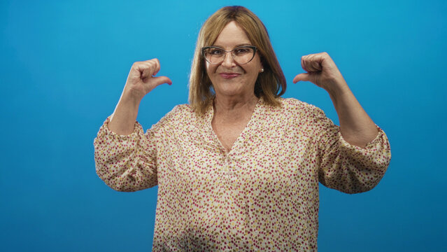 Middle age woman wearing glasses points both thumbs to her chest in a blue studio; confidence empowerment.
