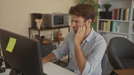 Man at desktop computer wearing striped shirt with hand on cheek and toothache in office building near bookshelves and plant  discomfort concentration. © Krakenimages.com