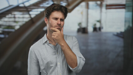 Naklejka premium Man touching chin and pursing lips in a modern building atrium, leaning near staircase and wearing striped shirt; pensive thought.