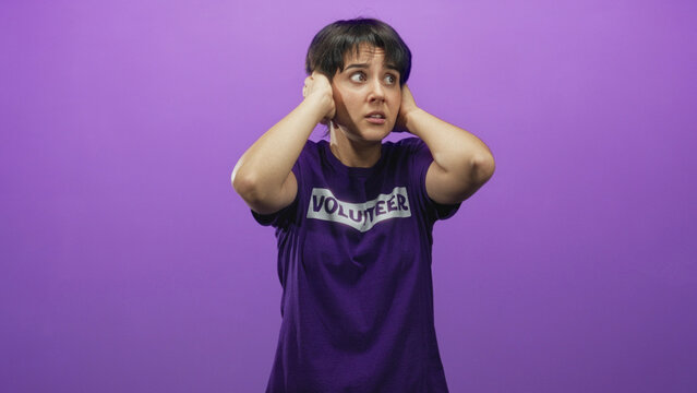 Woman volunteer in purple shirt covers ears with hands and grimaces in a studio while flinching from noise and looking distressed; anxiety overload.