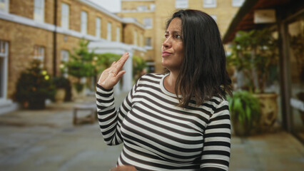 Fototapeta premium Woman with brunette hair in striped shirt raises hand palm outward in a dismissive gesture and touches chest with other hand in a brick courtyard building; skepticism doubt.