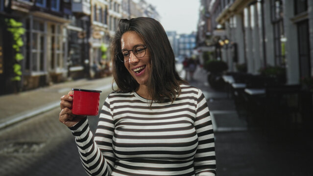 Woman holding a red enamel mug, smiling and tilting it toward camera while standing on a narrow cobblestone street outside a sidewalk cafe; contentment leisure.