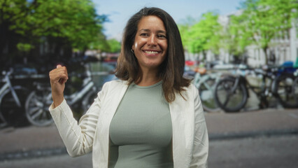 Fototapeta premium Brunette middle age woman smiling and giving a thumbs up while pointing at parked bicycles on a street lined with trees; confidence travel.