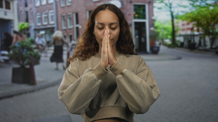 Fototapeta premium Woman with hands pressed together in prayer on a cobblestone street, cropped sweatshirt visible; serenity reflection.