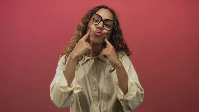 Woman presses fingers to cheeks in studio with pink backdrop and oversized glasses, pouting expression; playful curiosity.