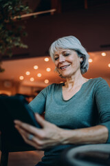 Smiling senior woman using tablet in cozy café. Scene radiates digital ease, joyful learning, and empowerment through personal tech in relaxed environment. © BullRun