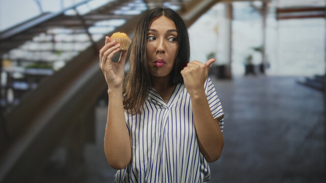 Hispanic brunette woman holding a muffin in one hand while thumb pointing to the side with the other, standing near an escalator in a building; skepticism reluctance.