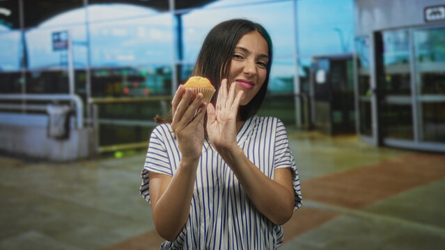 Young hispanic woman in striped shirt holds a muffin in one hand and shows palm away with the other hand at an airport terminal entrance while grimacing at the pastry; disgust refusal.