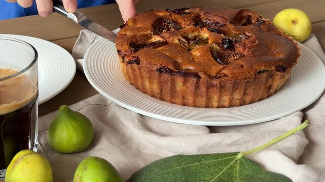 Rustic fruit tart on a white plate as hands slice it with a knife, surrounded by fresh figs and pears on a wooden table, warm homemade dessert mood