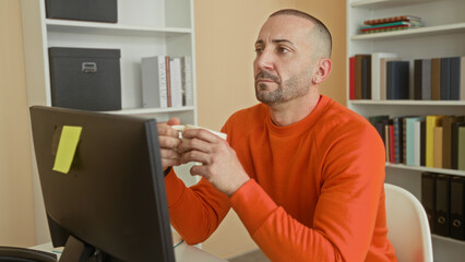 Man in orange sweater holds mug with both hands at computer desk among bookshelves in building, gazing at screen  quiet contemplation. © Krakenimages.com