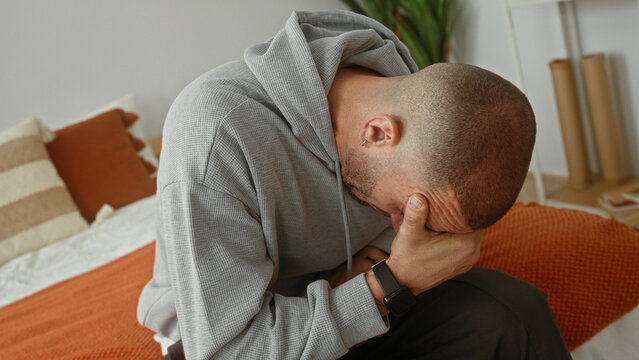 Man on bed sitting covers face with hand in a building bedroom, gray hoodie and watch on wrist visible; sadness fatigue.
