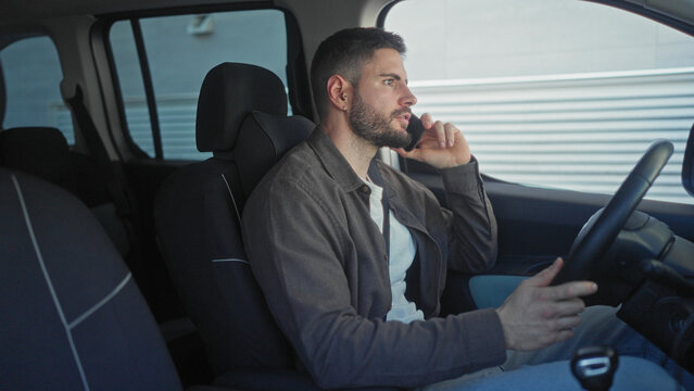 Man holding smartphone to ear while seated at steering wheel inside a car beside an industrial building, talking and gesturing with his free hand; concern.