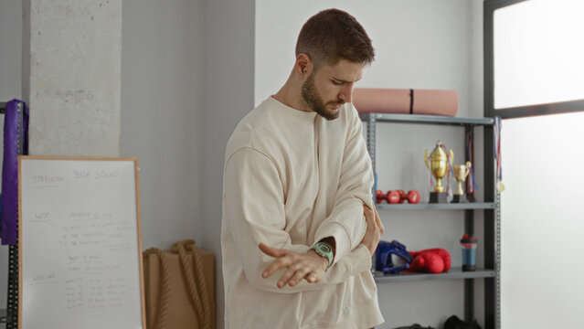 Man adjusting wristwatch and stretching forearm while rubbing elbow near shelves with boxing gloves and trophies in a training studio; focus preparation.