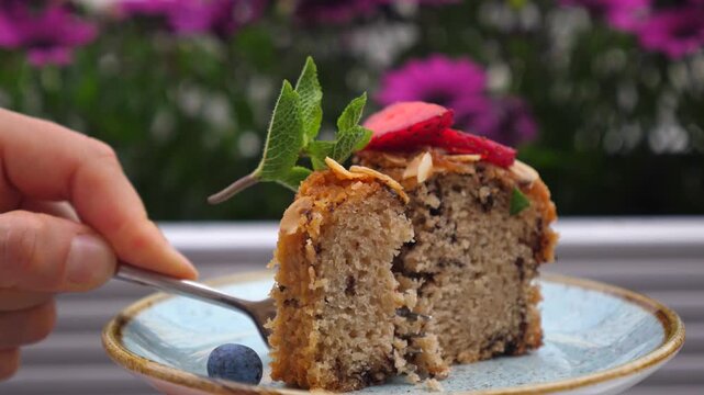 Close up view of a person using a fork to cut a delectable slice of homemade cake, garnished with fresh mint leaves, sliced strawberries, almonds, and a single blueberry on the side
