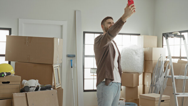 Man takes selfie with smartphone amid packed moving boxes, ladder, helmet and bubblewrap in building while unpacking and organizing; optimism new beginning.