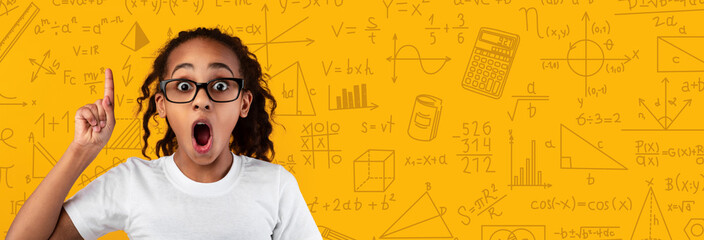 A young girl with curly hair and glasses looks surprised while raising her finger in a classroom. She stands against a bright background filled with math and science symbols. © Prostock-studio