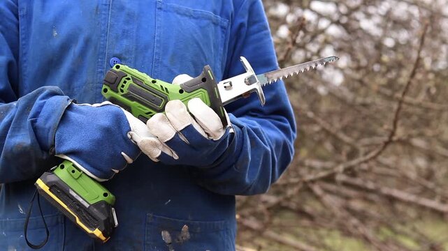 A worker in blue overalls holds a cordless reciprocating saw. A craftsman in protective gloves demonstrates a compact cordless reciprocating saw (hacksaw) in green. Professional power tool.