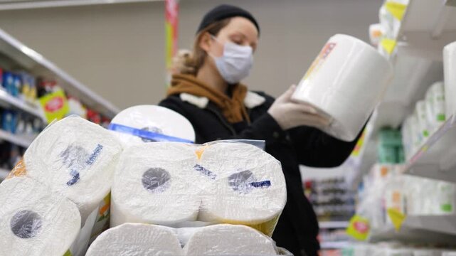 Scared woman wearing a protective face mask and gloves shopping for toilet paper at the supermarket, stocking up on supplies and bulk buying goods during the coronavirus pandemic quarantine