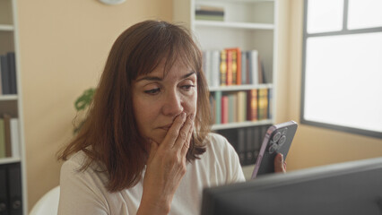 Fototapeta premium Woman holding smartphone, finger to cheek and phone near lips while checking screen in office building; privacy concern.