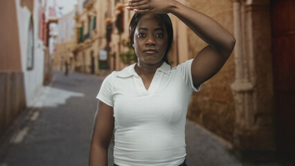 Obraz premium Woman in white polo holding hand to forehead, shading eyes while standing in a narrow cobblestone street lined with old stone buildings; heat fatigue.