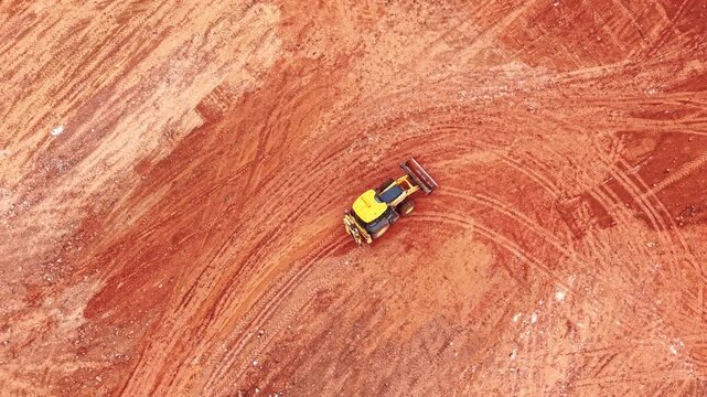 Top down drone view of industrial backhoe on red ground. Yellow excavator performing earthworks on red dry soil with circular tire patterns.
