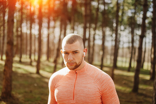 Man runs in the forest wearing earbuds and orange workout shirt while trees surround him during late afternoon sunlight