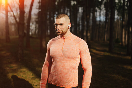 Man in orange shirt listens to music while standing in a forest during daylight with tall trees around him
