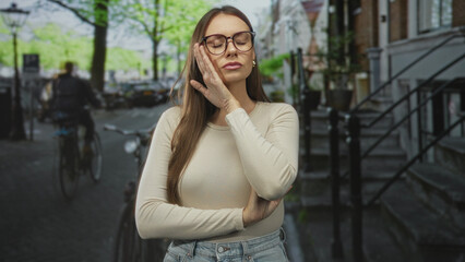 Fototapeta premium Woman with hand to cheek and crossed arm wearing glasses on street with bicycles and stone steps; pensive contemplation.