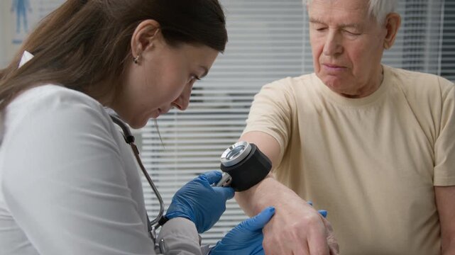 Young adult female medical professional wearing protective gloves inspecting forearm of elderly man within dermatoscope during routine lesion examination at modern clinic