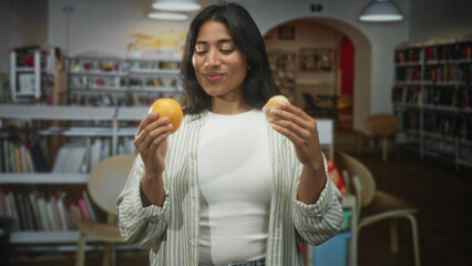 Young hispanic woman holding an orange and a muffin in her hands, smiling and comparing the snacks while standing among bookshelves in a public library building  choice contemplation. © Krakenimages.com