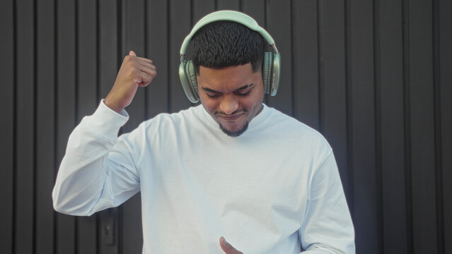 Man wearing headphones gives thumbs up and fist pump while nodding head against a building wall; joy groove.