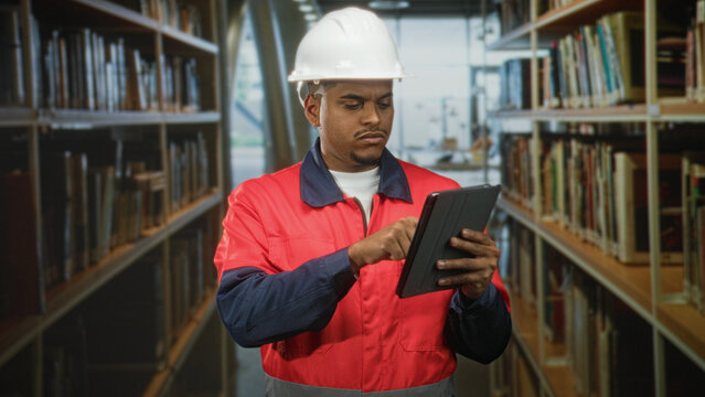Hispanic man construction worker with beard wearing red jacket and hard hat taps tablet with finger among library shelves inside a building; safety focus.