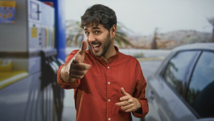 Smiling young hispanic man in red shirt points finger to camera near gas pump and car on street  playful confidence. © Krakenimages.com