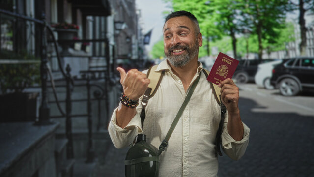 Man holding passport with thumb raised on street, backpacker wearing backpack and canteen, smiling while pointing thumb to passport; adventure travel joy.